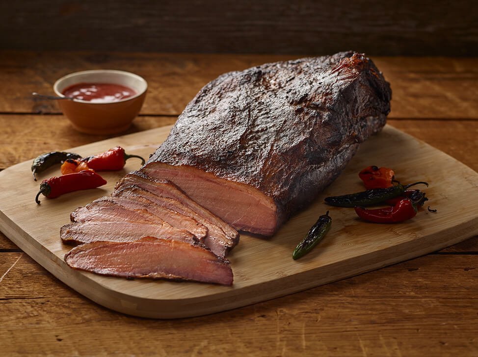 A side view of SADLER'S SMOKEHOUSE® whole brisket on a cutting board with some chilies and a ramekin or bbq sauce in the background.