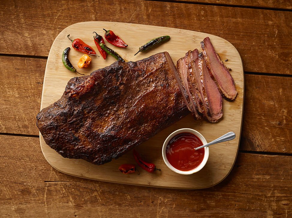 A top view of SADLER'S SMOKEHOUSE® whole brisket on a cutting board with some chilies and a ramekin or bbq sauce.