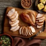 A carved SADLER'S SMOKEHOUSE® whole turkey on a cutting board with sides of corn bread, salad, and baked beans in the background.