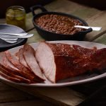 SADLER'S SMOKEHOUSE® turkey breast on a white serving dish with a cast iron of backed beans behind it.