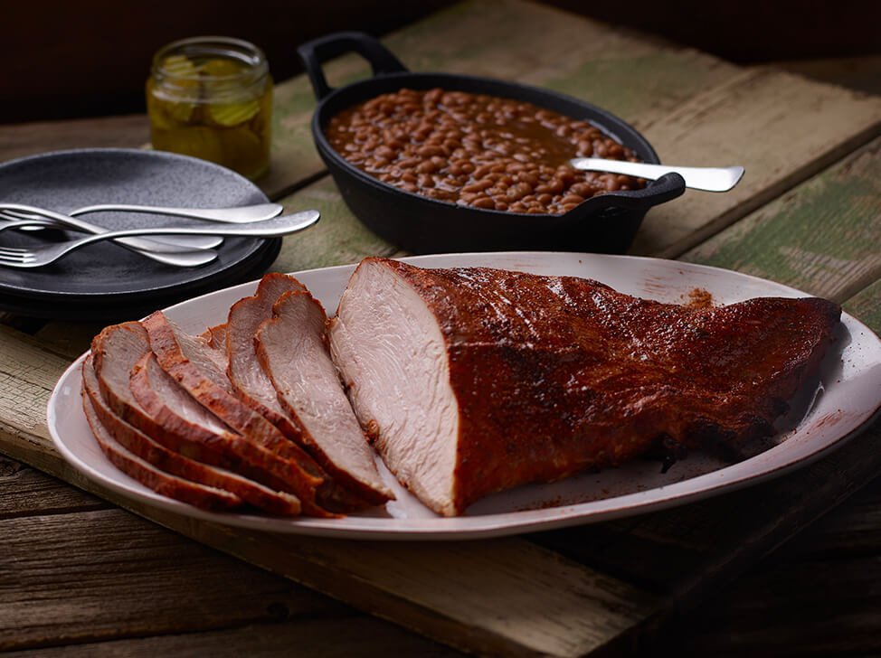SADLER'S SMOKEHOUSE® turkey breast on a white serving dish with a cast iron of backed beans behind it.