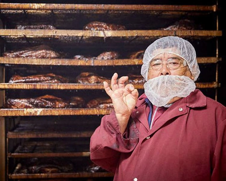 Pit master standing in front of racks of bbq meat making the "okay" symbol with his hand.